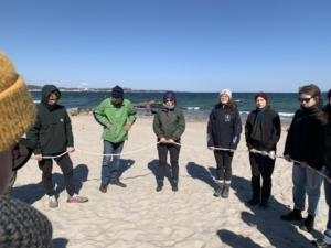 Gruppe von jungen Erwachsenen die an einem Strand im Kreis stehen und ein Seil festhalten.