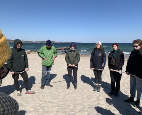 Gruppe von jungen Erwachsenen die an einem Strand im Kreis stehen und ein Seil festhalten.