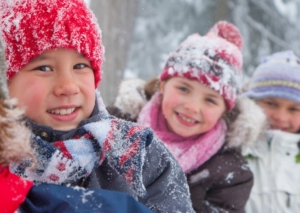 Drei Kinder im winterlichen Wald. Sie tragen verschneite Winterkleidung.