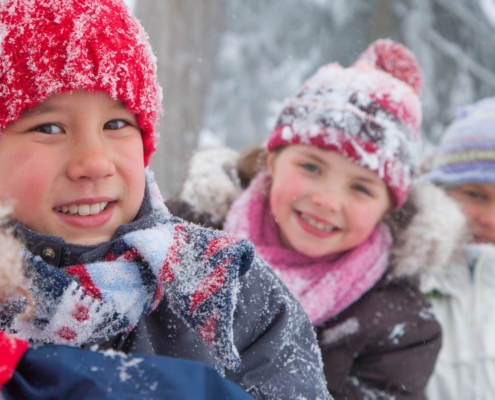 Drei Kinder im winterlichen Wald. Sie tragen verschneite Winterkleidung.