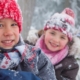 Drei Kinder im winterlichen Wald. Sie tragen verschneite Winterkleidung.