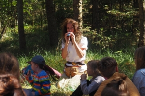 Frau sitzt mit Gruppe von Kindern im Wald. Sie pfeift durch eine Pfeife.