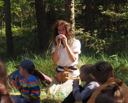 Frau sitzt mit Gruppe von Kindern im Wald. Sie pfeift durch eine Pfeife.